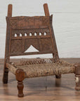 Brown wooden low chair with rope seat and weathered finish on a wooden floor, next to a white brick wall indoors.