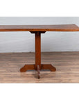 Brown wooden Indonesian console table with single plank top and pedestal base on wood floor against white brick wall.