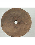 Brown wooden ox cart wheel on black base, displayed indoors in front of a light brick wall.