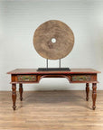 Brown wooden ox cart wheel on black painted base, rustic style, displayed behind an antique table indoors.