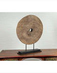 Brown wooden ox cart wheel on black base, displayed on a table with white brick wall background.
