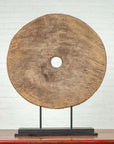 Brown wooden ox cart wheel from Thailand on a black base, displayed indoors against a white brick wall.