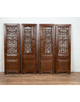 Natural elm wood carved screen panels with fretwork, foliage, and floral motifs against a white brick wall.