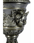 Close-up of a silver plate urn with cherub face and ornate leaf relief designs.