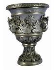 Silver plate urn with embossed leaves, faces, and cherub ornaments on base and body, close-up.