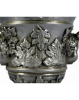 Close-up of silver plate urn with ornate leaf patterns, sculpted faces, and figural cherub ornaments.
