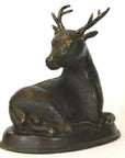 Bronze sitting deer sculpture with antlers on an oval base, close-up against a white background.