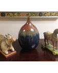 Close-up of a brown and blue ceramic Prem vase on a wooden surface, between wheeled cow and horse figurines.