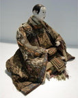 Close-up of a Taisho Samurai Doll seated with hands on lap, wearing ornate patterned robes.