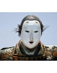 Close-up of a Taisho samurai doll with a white Noh mask, black hair tufts, and ornate patterned clothing.