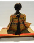 Black-haired Taisho Samurai Doll in floral kimono sitting on woven mat, rear view.