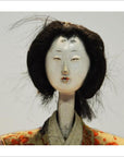 Close-up of Taisho Samurai Doll with white face, black hair, and patterned kimono against a plain background.
