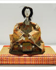 Close-up of Taisho Samurai Doll with painted face and ornate kimono on a mat with a red patterned border.