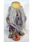 Gray-haired doll with embroidered clothing and worn beige headpiece, close-up, indoors.