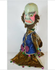 Blue and gold fabric doll with green face, bold features, and pink bow, close-up.