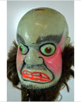 Close-up of painted Taiwan opera doll mask with green eyes, red cheeks and mouth, bushy brows, dark brown hair tufts.