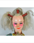 Close-up of Taiwan Opera Doll with gray pigtails, bright pink makeup, and painted smile against plain background.
