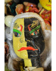 Yellow and black hand-painted mask with green brows, white hair, and red lips, ceramic close-up on plain background.