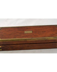 Brown teakwood pencil and jewelry box with brass accents and nameplate, closed, on a light background.