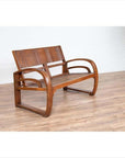 Brown teak wood settee with folding back, looping arms, and cane seat on light wooden floor indoors against white wall.