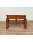 Brown teak wood settee with folding back, looping arms, and cane seat against a white brick wall indoors.