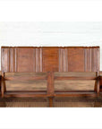 Brown teak wood settee with folding back, looping arms, and cane seat against a white brick wall indoors.