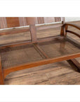 Brown teak wood settee with folding back, looping arms, and cane seat on wooden floor indoors.