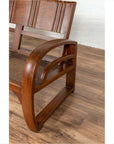Close-up of brown teak wood settee with cane seat, folding back, and looping arms on a wooden floor.