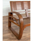 Brown teak wood settee with looping arms, folding back, and cane seat on a wooden floor indoors.