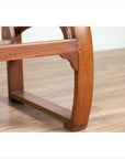 Close-up of brown teak wood settee with looping arms, folding back, and cane seat on a light wood floor indoors.