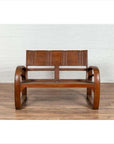 Brown teak wood settee with looping arms, folding back, and cane seat on a wooden floor indoors.