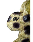 Close-up of ceramic Thai Begging Rabbit figurine with brown spots and red eye on a white background, facing left.
