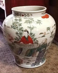 White porcelain urn with painted figures, bamboo, and outdoor scene on a carpeted floor near wooden furniture.