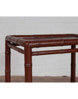 Brown bamboo side table with recessed top, vintage 1950s, against white brick wall and wood floor indoors.