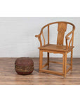 Brown rattan circular hat box with weathered patina beside a wooden armchair on a wood floor indoors.