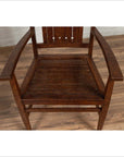 Brown wooden Dutch Colonial armchair with pierced slats and bonnet-style rail, on wood floor by white brick wall indoors.