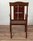 Brown wooden Dutch Colonial armchair with pierced slats and bonnet rail on wooden floor by white brick wall indoors.