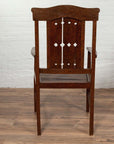 Brown wooden Dutch Colonial armchair with pierced slats and bonnet rail on wooden floor by white brick wall indoors.