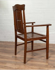 Brown wood Dutch Colonial armchair with pierced slats and bonnet rail on wooden floor near white brick wall indoors.