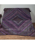 Purple silk fabric with silver and gold accents displayed on a surface next to a wooden floor and white brick wall.