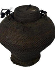 Brown woven basket made from natural fibers with wide body, narrow base, fitted lid, and braided handles.
