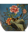 Close-up of a vintage porcelain vase with hand painted pink flowers and a bird on a rock, set against a blue background.