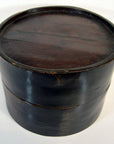 Close-up of round dark brown wooden hat box with Chinese design, lid on, light and dark wood, visible scratches.