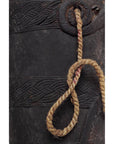 Close-up of a braided rope looped through a decorative brown wooden Indian pitcher on a plain background.