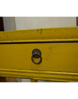 Close-up of yellow wood side table drawer with distressed finish and round metal ring pull handle at center.
