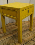 Yellow wood side table with single drawer and horse hoof feet, on wooden floor indoors.