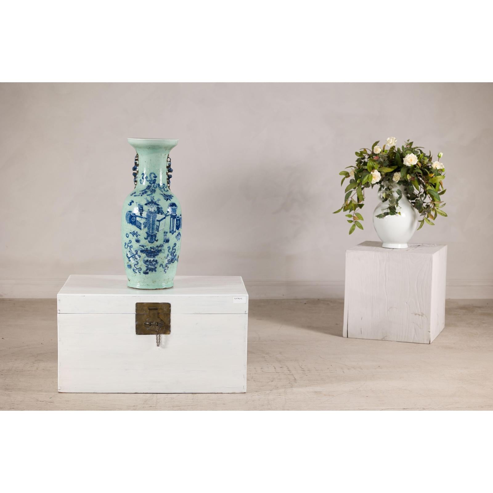 Cobalt blue porcelain vase with scholar images on wooden chest, white floral vase on pedestal in background.