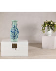 Cobalt blue porcelain vase with scholar images on wooden chest, white floral vase on pedestal in background.