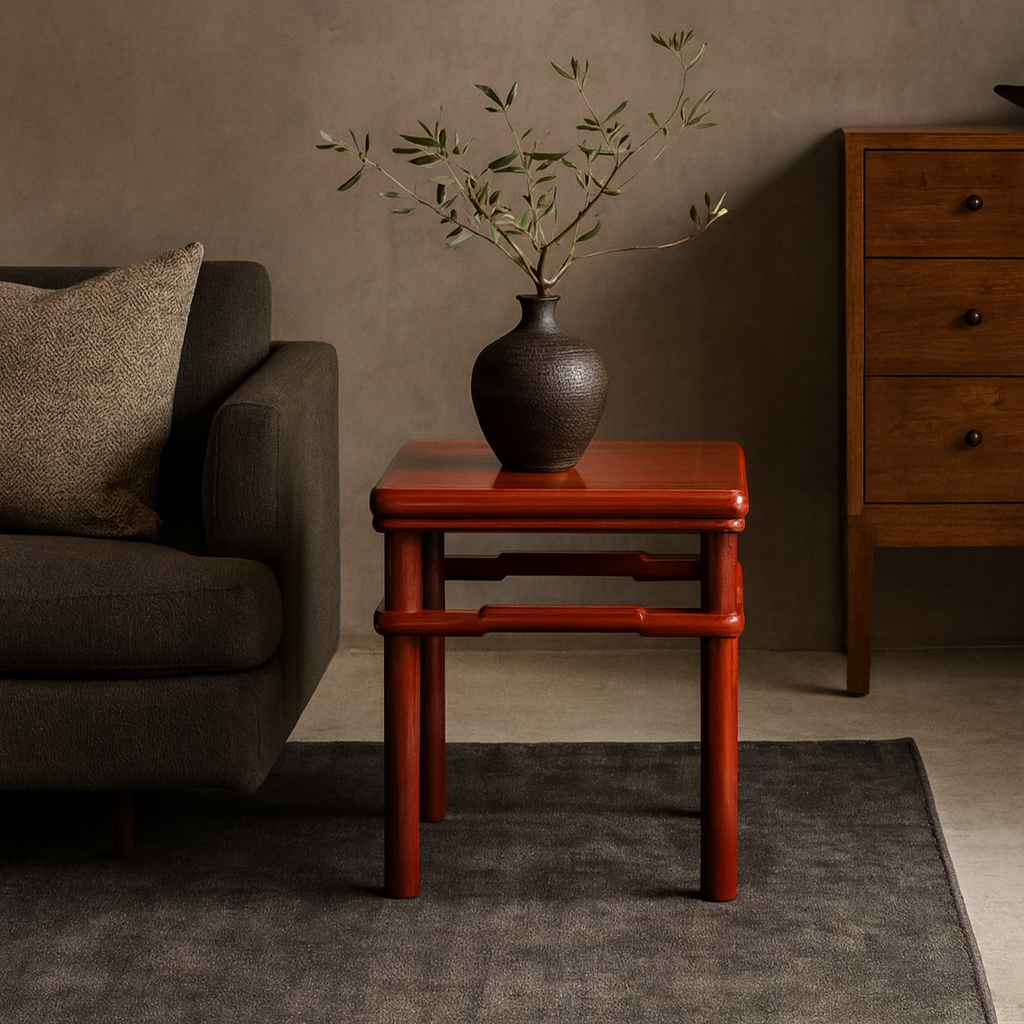 Dark ceramic vase with leafy branches on red square side table beside gray sofa and wooden dresser.