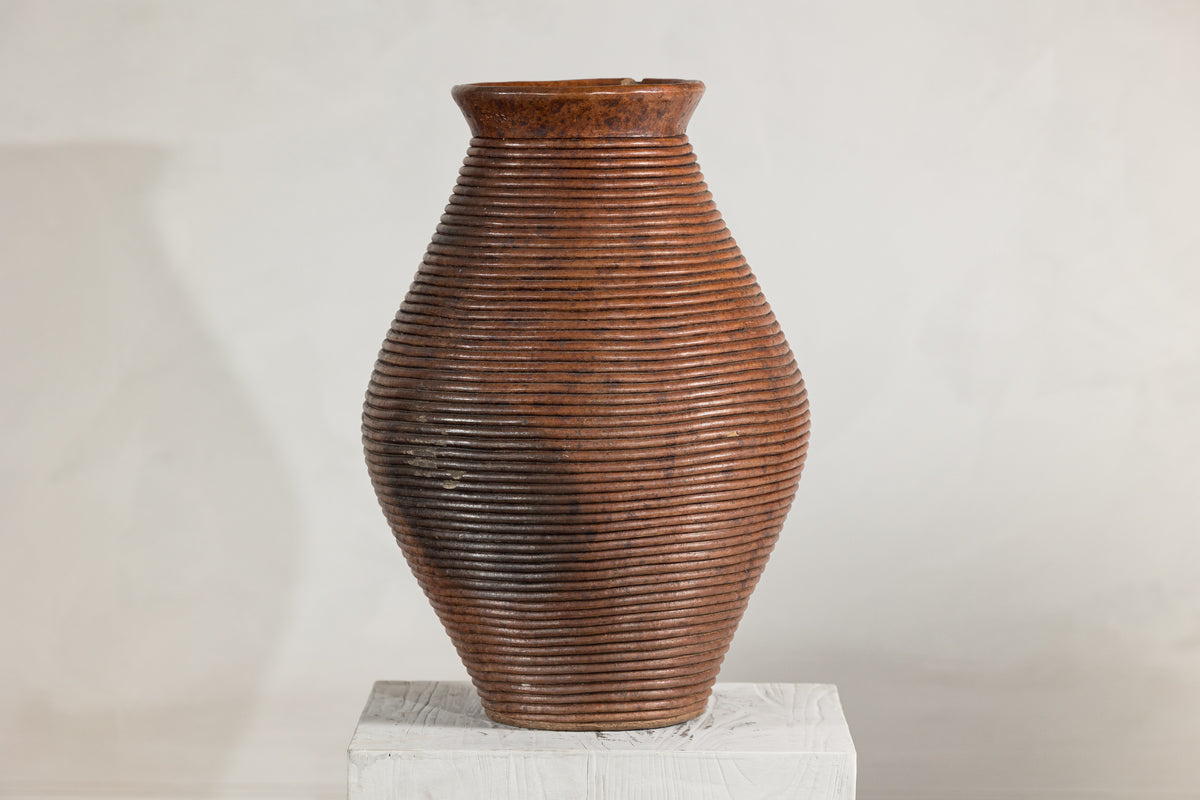 Brown ribbed pottery vase with tall narrow neck and burnished finish on white pedestal against plain background.